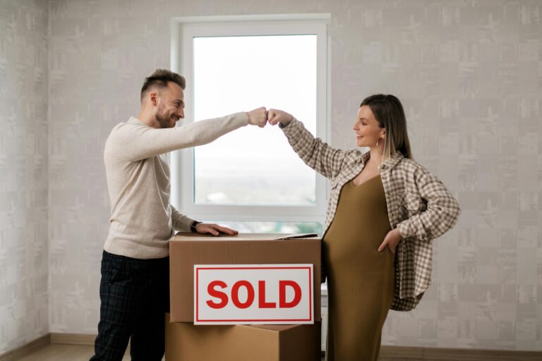 Pregnant couple celebrating new home purchase with boxes labeled 'SOLD'.